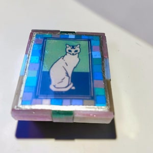 May include: A rectangular box featuring a central illustration of a white cat sitting against a green and blue background. The lid is bordered by a mosaic pattern of small blue, purple, and light blue tiles, framed by a metallic edge finish.