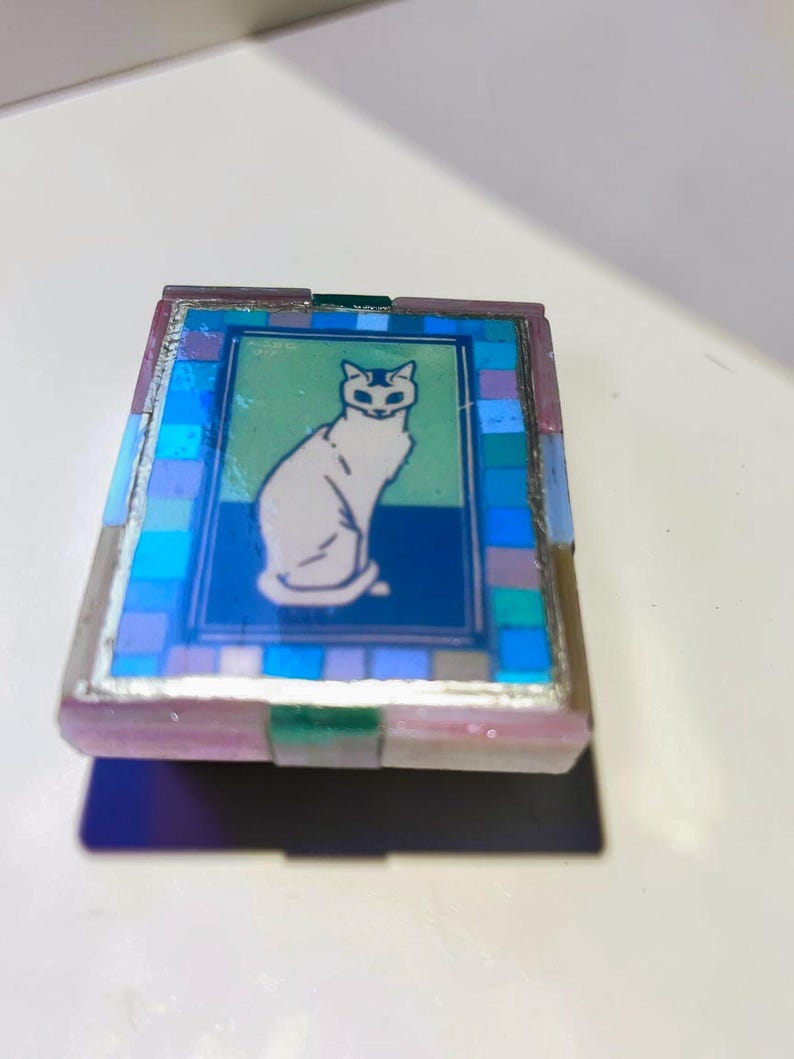 May include: A rectangular box featuring a central illustration of a white cat sitting against a green and blue background. The lid is bordered by a mosaic pattern of small blue, purple, and light blue tiles, framed by a metallic edge finish.