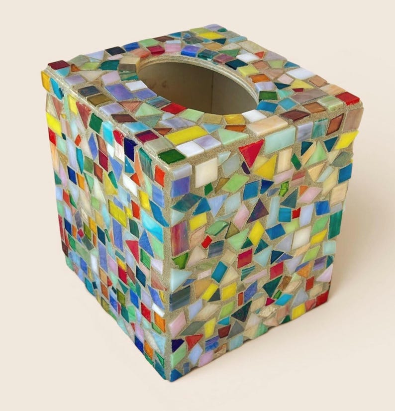 May include: A square tissue box cover featuring a mosaic design made of small, multi-colored glass tiles in shades of red, blue, yellow, green, and white. The tiles are arranged in a random pattern and set into a light-colored grout base.