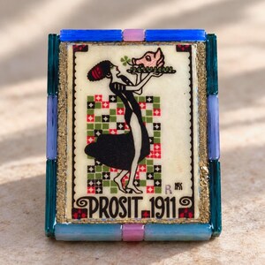 May include: A rectangular decorative tile featuring a central illustration of a figure holding a platter with a pig head. The text PROSIT 1911 is printed at the bottom. The tile is framed by blue, teal, and pink glass-like rectangular beads.