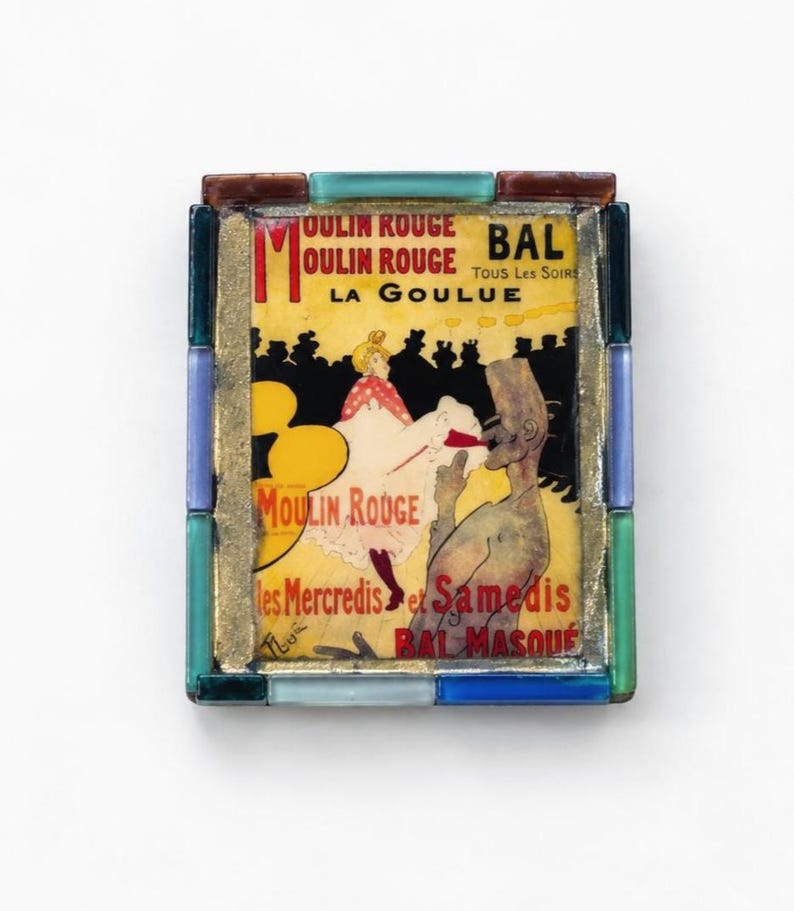 May include: A rectangular decorative tile featuring a Moulin Rouge La Goulue poster print, framed by a border of small, multi-colored glass tiles in shades of blue, green, and brown, with gold-toned metal accents surrounding the central image.