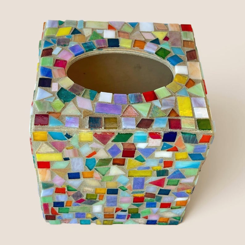 May include: A square tissue box cover featuring a multi-colored mosaic design made of small, irregular glass tiles in shades of red, blue, yellow, green, and purple, set in light-colored grout with a large oval opening at the top center.