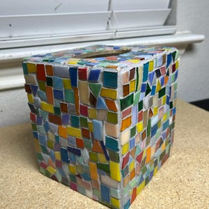 May include: A square tissue box cover featuring a multi-colored mosaic design. The exterior is composed of numerous small, irregularly shaped glass tiles in shades of red, blue, yellow, green, orange, and white, all set within light gray grout.