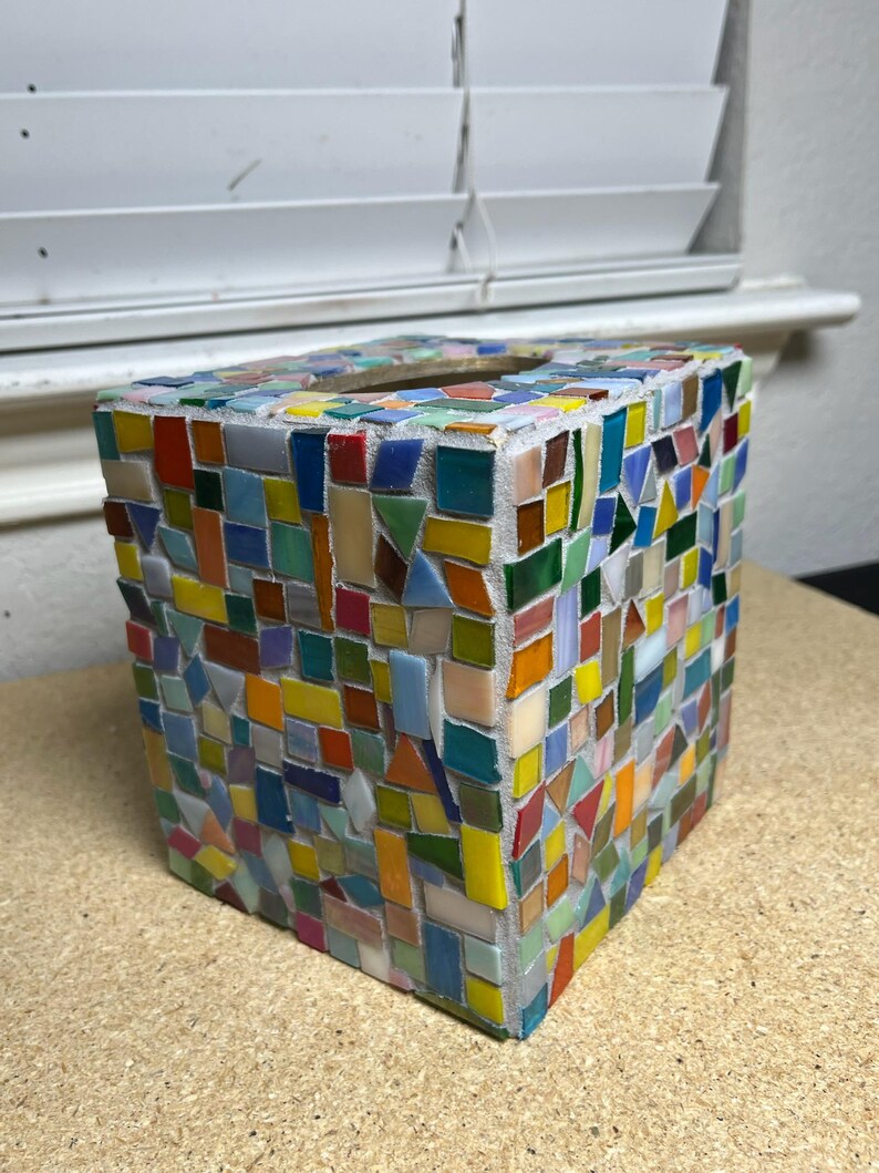 May include: A square tissue box cover featuring a multi-colored mosaic design. The exterior is composed of numerous small, irregularly shaped glass tiles in shades of red, blue, yellow, green, orange, and white, all set within light gray grout.