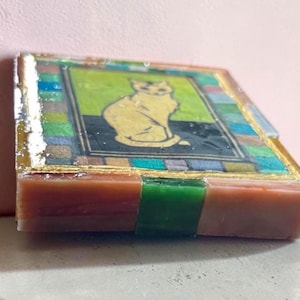 May include: A square coaster featuring a central illustration of a seated cat against a lime green and black background. The border consists of a mosaic-style pattern of multicolored squares in blue, green, and brown, finished with a glossy resin coating.