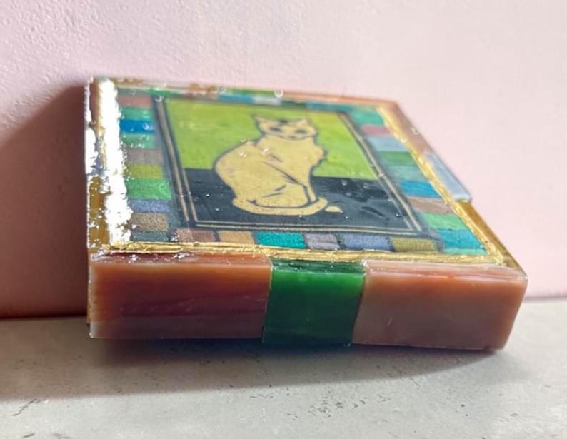 May include: A square coaster featuring a central illustration of a seated cat against a lime green and black background. The border consists of a mosaic-style pattern of multicolored squares in blue, green, and brown, finished with a glossy resin coating.
