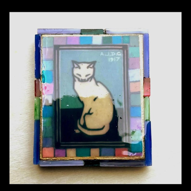 May include: A rectangular decorative tile featuring a central illustration of a seated cat in cream and white tones against a dark background. The text A.J.D.G 1917 is visible. The center is framed by a border of small, multicolored glass mosaic squares.