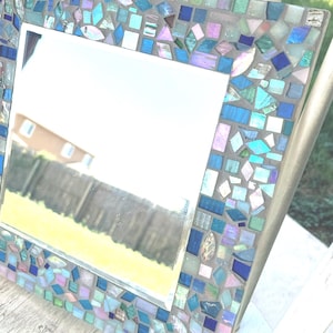 Op de afbeelding: Een vierkante spiegel met een decoratieve lijst bestaande uit kleine, onregelmatig gevormde moza&iuml;ektegels in tinten blauw, groenblauw, groen en iriserend roze, gezet in een lichtgrijze voegbasis met een reflecterend glazen middengedeelte.