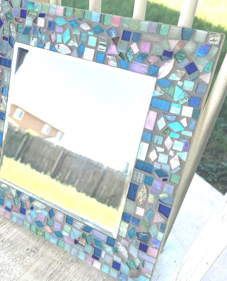 Op de afbeelding: Een vierkante spiegel met een decoratieve lijst bestaande uit kleine, onregelmatig gevormde moza&iuml;ektegels in tinten blauw, groenblauw, groen en iriserend roze, gezet in een lichtgrijze voegbasis met een reflecterend glazen middengedeelte.