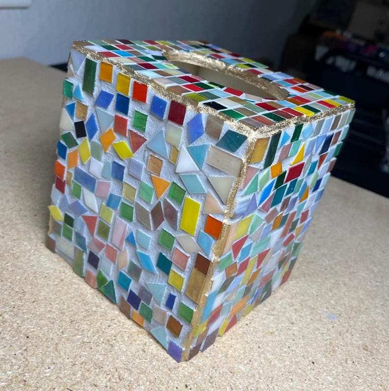 K&ouml;nnte beinhalten: Ein quadratischer Taschentuchbox-Bezug mit einem bunten Mosaikmuster aus kleinen, unregelm&auml;&szlig;ig geformten Glasfliesen in Rot, Blau, Gr&uuml;n, Gelb und Orange, mit goldfarbenen Kanten und einer ovalen &Ouml;ffnung an der Oberseite des Objekts.