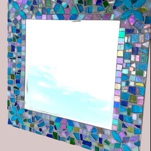 Puede incluir: Un espejo de pared cuadrado con un marco de mosaico decorativo compuesto por varias piezas de vidrio en tonos azul, turquesa, morado y verde. El marco incluye cuatro acentos distintos en forma de flor hechos de p&eacute;talos de vidrio turquesa brillante.