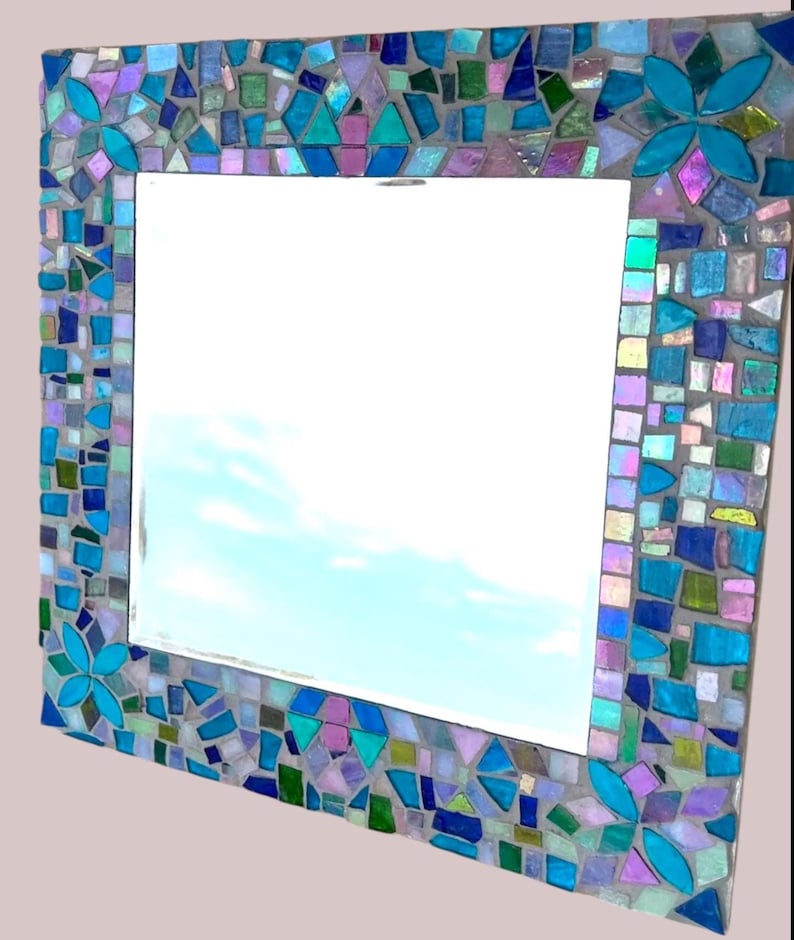 Puede incluir: Un espejo de pared cuadrado con un marco de mosaico decorativo compuesto por varias piezas de vidrio en tonos azul, turquesa, morado y verde. El marco incluye cuatro acentos distintos en forma de flor hechos de p&eacute;talos de vidrio turquesa brillante.