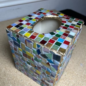K&ouml;nnte beinhalten: Ein quadratischer Taschentuchbox-Bezug, verziert mit einem bunten Mosaikmuster aus kleinen Glasfliesen in Rot-, Blau-, Gr&uuml;n-, Gelb- und Wei&szlig;t&ouml;nen. Die obere &Ouml;ffnung ist mit einem strukturierten goldfarbenen Rand versehen und die Fliesen sind in wei&szlig;em Fugenm&ouml;rtel gesetzt.