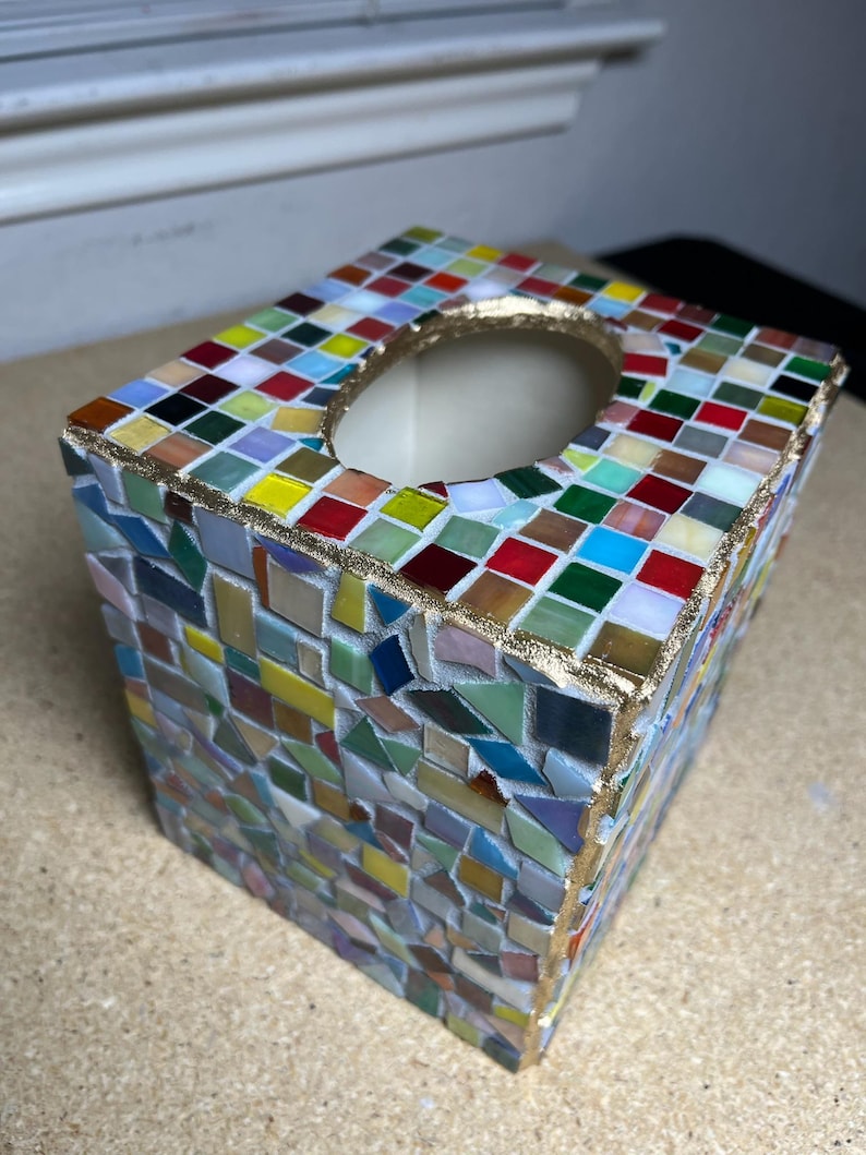 K&ouml;nnte beinhalten: Ein quadratischer Taschentuchbox-Bezug, verziert mit einem bunten Mosaikmuster aus kleinen Glasfliesen in Rot-, Blau-, Gr&uuml;n-, Gelb- und Wei&szlig;t&ouml;nen. Die obere &Ouml;ffnung ist mit einem strukturierten goldfarbenen Rand versehen und die Fliesen sind in wei&szlig;em Fugenm&ouml;rtel gesetzt.
