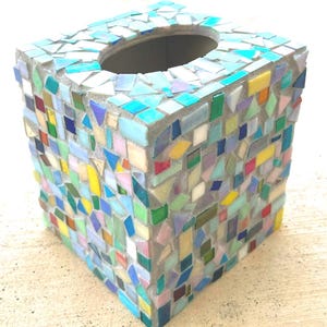 May include: A square tissue box cover decorated with a mosaic of small, multi-colored glass tiles in shades of blue, green, yellow, pink, and white. The tiles are set in gray grout, forming a geometric pattern across the sides and top surface.