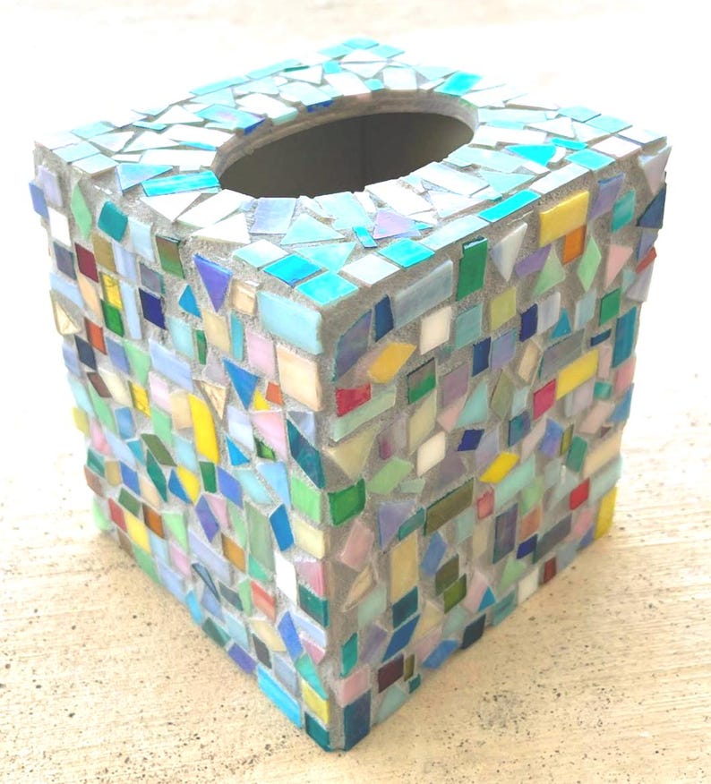 May include: A square tissue box cover decorated with a mosaic of small, multi-colored glass tiles in shades of blue, green, yellow, pink, and white. The tiles are set in gray grout, forming a geometric pattern across the sides and top surface.
