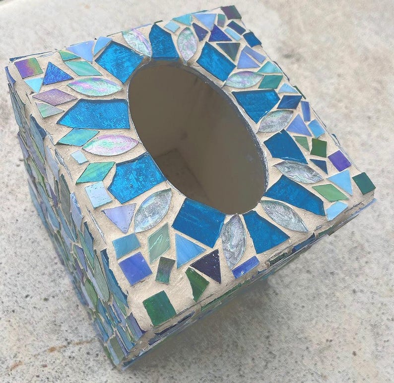 Op de afbeelding: Een vierkante tissueboxhouder versierd met een moza&iuml;ek van blauwe, groene en iriserende glasstukjes in lichtgekleurde voegspecie. De bovenkant heeft een ovale opening en de zijkanten zijn bedekt met diverse geometrische glassplinters.