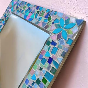 Puede incluir: Un espejo cuadrado con un marco de mosaico compuesto por varias piezas de vidrio en tonos turquesa, azul cobalto, verde claro y rosa suave. Las piezas est&aacute;n dispuestas en un patr&oacute;n geom&eacute;trico con un dise&ntilde;o de flor de cuatro p&eacute;talos.