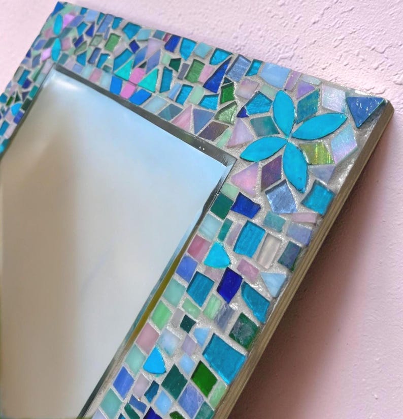Puede incluir: Un espejo cuadrado con un marco de mosaico compuesto por varias piezas de vidrio en tonos turquesa, azul cobalto, verde claro y rosa suave. Las piezas est&aacute;n dispuestas en un patr&oacute;n geom&eacute;trico con un dise&ntilde;o de flor de cuatro p&eacute;talos.