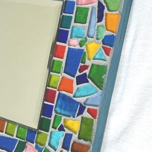 May include: A rectangular wall mirror featuring a mosaic frame constructed from numerous small, irregularly shaped pieces of translucent glass in vibrant shades of red, orange, yellow, green, and blue, all set within a light gray grout border.