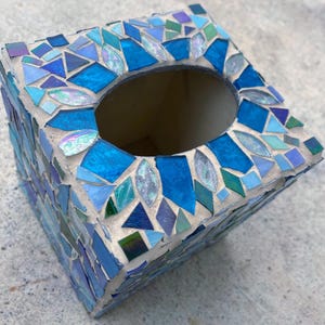 Op de afbeelding: Een vierkante tissueboxhouder versierd met een moza&iuml;ek van blauwe, groenblauwe en iriserende glastegels in lichtgrijze voegmortel. De bovenkant heeft een centrale ovale opening en is bekleed met diverse geometrische glasvormen.