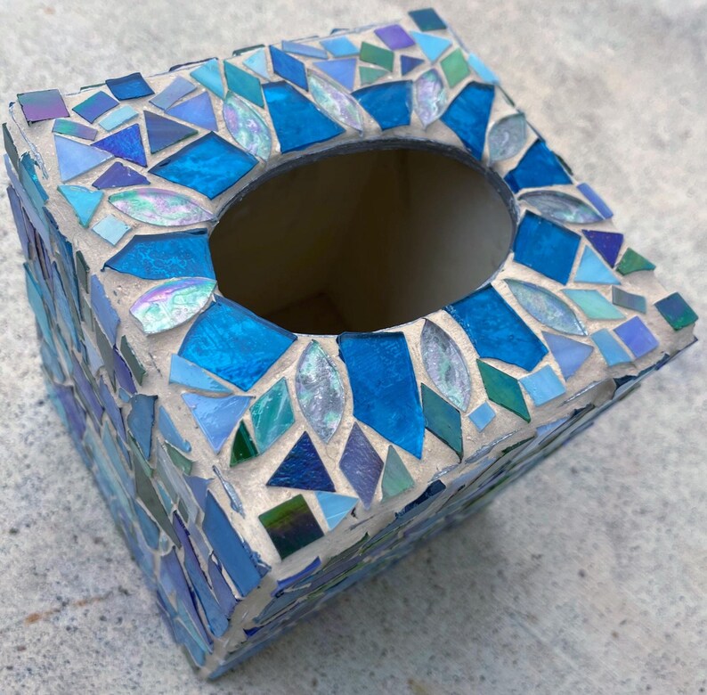 Op de afbeelding: Een vierkante tissueboxhouder versierd met een moza&iuml;ek van blauwe, groenblauwe en iriserende glastegels in lichtgrijze voegmortel. De bovenkant heeft een centrale ovale opening en is bekleed met diverse geometrische glasvormen.