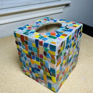 May include: A square tissue box cover decorated with a colorful mosaic pattern of small, irregularly shaped glass tiles in shades of blue, green, yellow, orange, and red, set into a light gray grout with an oval opening on the top surface.
