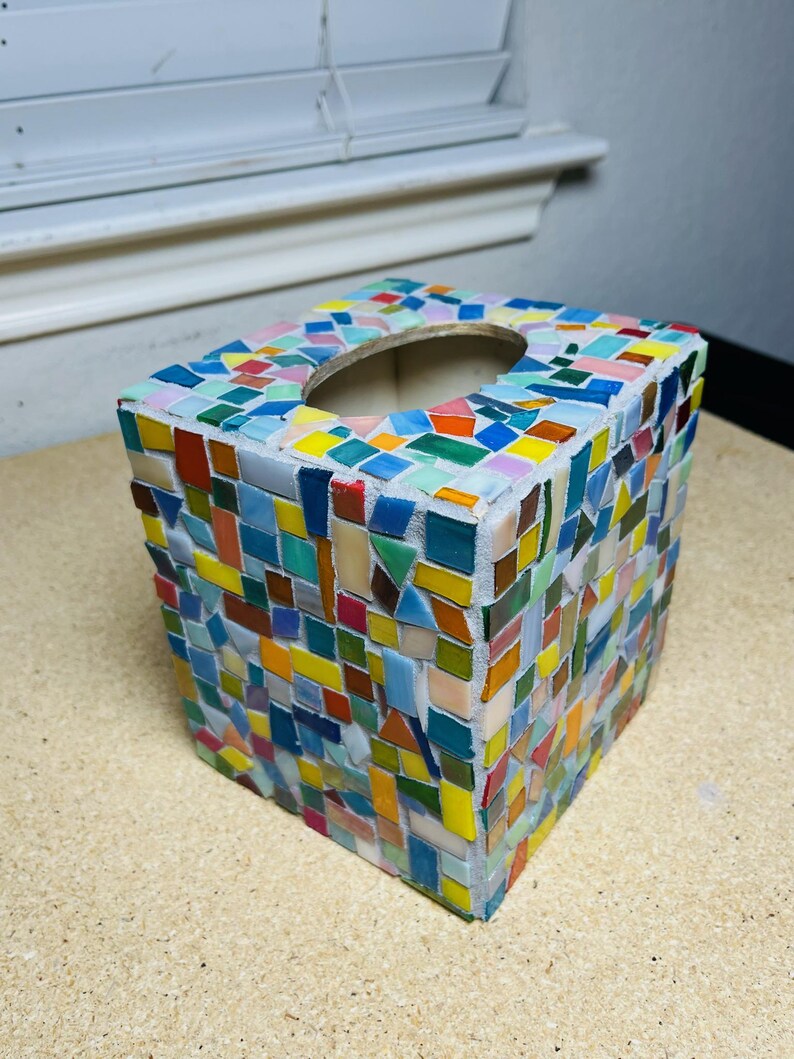 May include: A square tissue box cover decorated with a colorful mosaic pattern of small, irregularly shaped glass tiles in shades of blue, green, yellow, orange, and red, set into a light gray grout with an oval opening on the top surface.