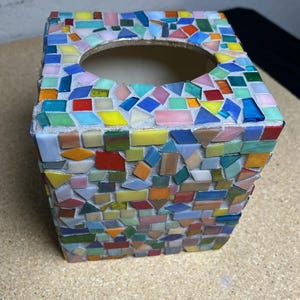 May include: A square tissue box cover featuring a mosaic design made of small, multi-colored glass tiles in shades of red, blue, yellow, green, and pink. The tiles are set in a light gray grout, creating a textured surface with a circular opening.