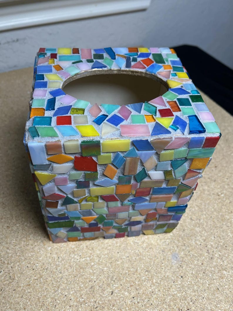May include: A square tissue box cover featuring a mosaic design made of small, multi-colored glass tiles in shades of red, blue, yellow, green, and pink. The tiles are set in a light gray grout, creating a textured surface with a circular opening.