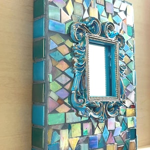 May include: A rectangular wall mirror featuring a mosaic border made of multi-colored glass tiles in shades of blue, green, yellow, and iridescent tones. A central ornate, scrolled frame in metallic blue and gold surrounds the reflective glass surface.