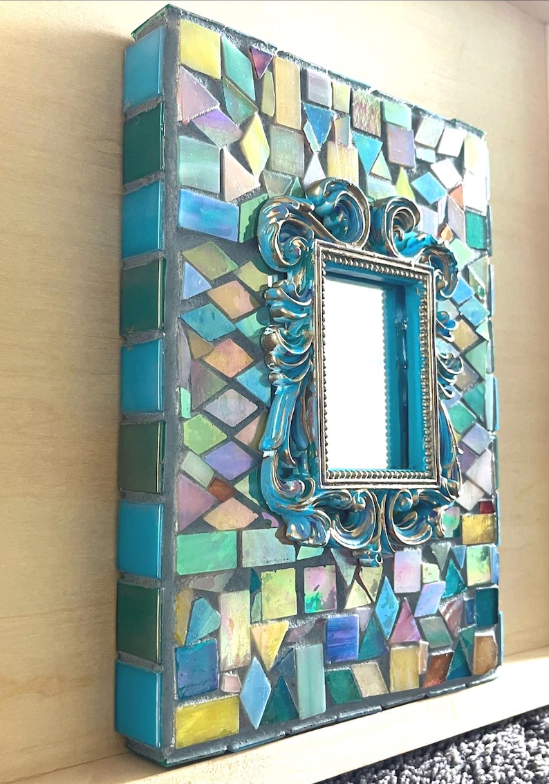May include: A rectangular wall mirror featuring a mosaic border made of multi-colored glass tiles in shades of blue, green, yellow, and iridescent tones. A central ornate, scrolled frame in metallic blue and gold surrounds the reflective glass surface.