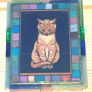 Pode incluir: Um azulejo decorativo retangular com uma ilustra&ccedil;&atilde;o central de um gato malhado bege sobre um fundo azul escuro com o texto AJDG 1918. O azulejo &eacute; emoldurado por uma borda de pequenos quadrados de mosaico de vidro multicoloridos.