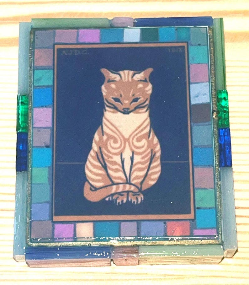 Pode incluir: Um azulejo decorativo retangular com uma ilustra&ccedil;&atilde;o central de um gato malhado bege sobre um fundo azul escuro com o texto AJDG 1918. O azulejo &eacute; emoldurado por uma borda de pequenos quadrados de mosaico de vidro multicoloridos.