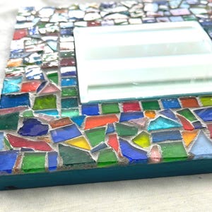 May include: A square mirror featuring a frame decorated with a mosaic of multicolored glass shards in shades of blue, green, red, orange, and yellow, set in gray grout. The side of the frame is painted a solid teal color, providing a contrast.