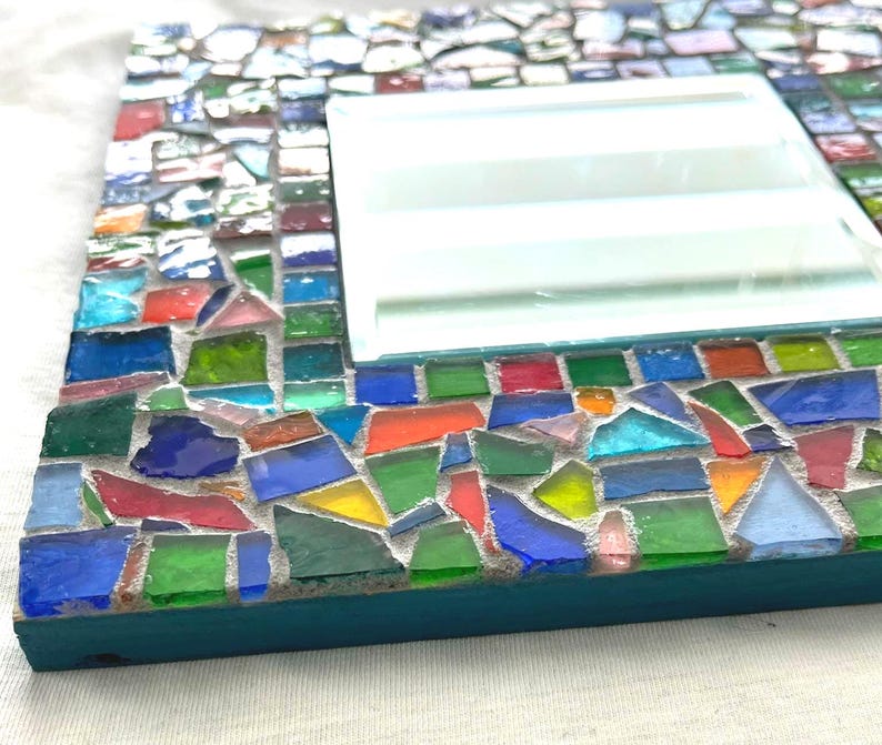 May include: A square mirror featuring a frame decorated with a mosaic of multicolored glass shards in shades of blue, green, red, orange, and yellow, set in gray grout. The side of the frame is painted a solid teal color, providing a contrast.