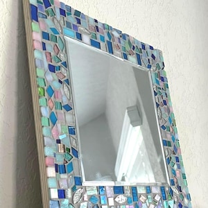Op de afbeelding: Een vierkante wandspiegel met een decoratieve moza&iuml;eklijst bestaande uit kleine, onregelmatig gevormde glastegels in tinten blauw, lichtroze, mintgroen en iriserend wit, geplaatst op een lichtgekleurd houten basisframe.
