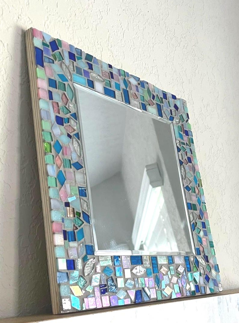 Op de afbeelding: Een vierkante wandspiegel met een decoratieve moza&iuml;eklijst bestaande uit kleine, onregelmatig gevormde glastegels in tinten blauw, lichtroze, mintgroen en iriserend wit, geplaatst op een lichtgekleurd houten basisframe.