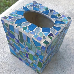 May include: A square tissue box cover decorated with a mosaic of glass pieces in various shades of blue, teal, and green. The pieces are arranged in an irregular pattern and set in light-colored grout, featuring an oval opening on the top surface.