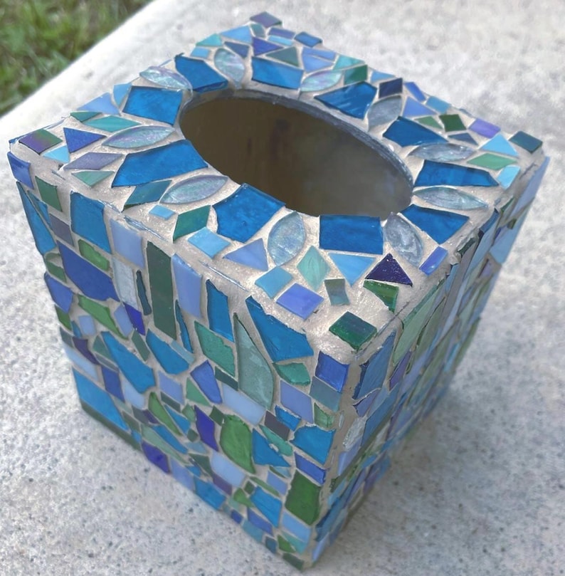 Op de afbeelding: Een vierkante tissueboxhouder versierd met een moza&iuml;ek van glasstukjes in diverse tinten blauw, groen en zeegroen. De stukjes zijn in een onregelmatig patroon geplaatst en vastgezet in lichtgekleurde voegspecie, met een ovale opening aan de bovenzijde.