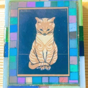 May include: A rectangular decorative tile featuring a central illustration of a seated orange tabby cat against a dark blue background. The text AJD.G. and 1913 are visible in the upper corners. The tile is framed by a border of small glass tiles.
