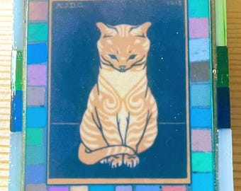 May include: A rectangular decorative tile featuring a central illustration of a seated orange tabby cat against a dark blue background. The text AJD.G. and 1913 are visible in the upper corners. The tile is framed by a border of small glass tiles.