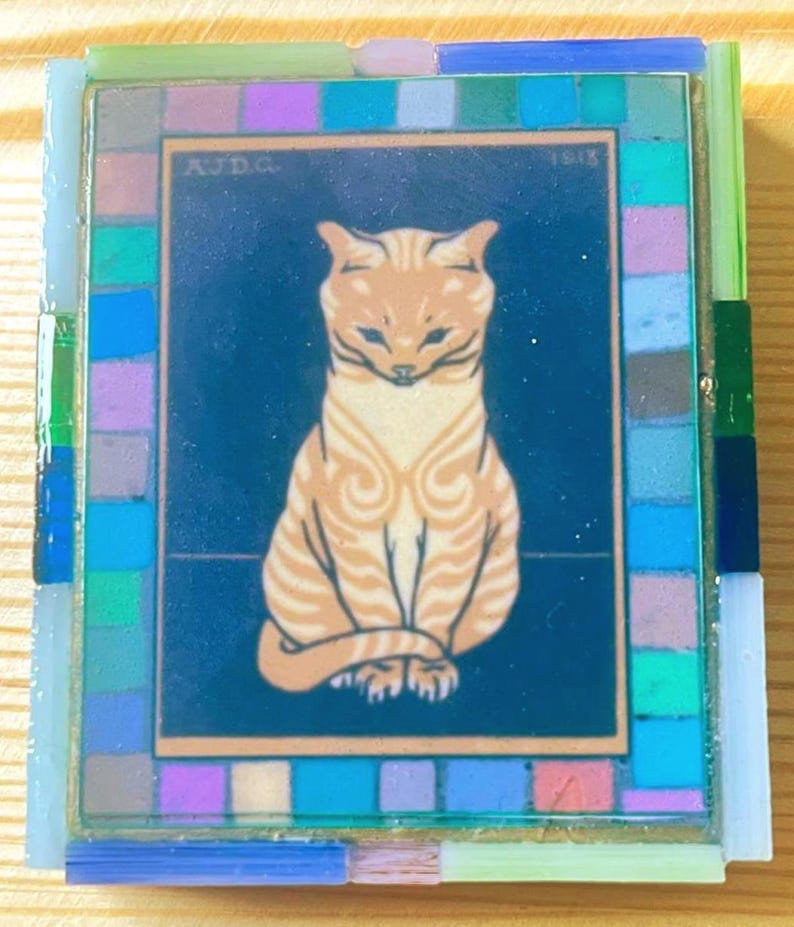 Pode incluir: Um azulejo decorativo retangular com uma ilustra&ccedil;&atilde;o central de um gato malhado laranja sentado sobre um fundo azul escuro. O texto AJD.G. e 1913 &eacute; vis&iacute;vel nos cantos superiores. O azulejo &eacute; emoldurado por uma borda de pequenas pe&ccedil;as de vidro.