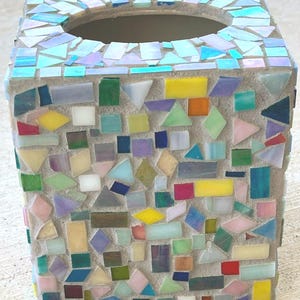 May include: A square tissue box cover decorated with a mosaic of multicolored glass tiles in shades of blue, yellow, green, pink, and orange. The tiles are set into a light gray grout, creating a textured surface with a circular opening on top.
