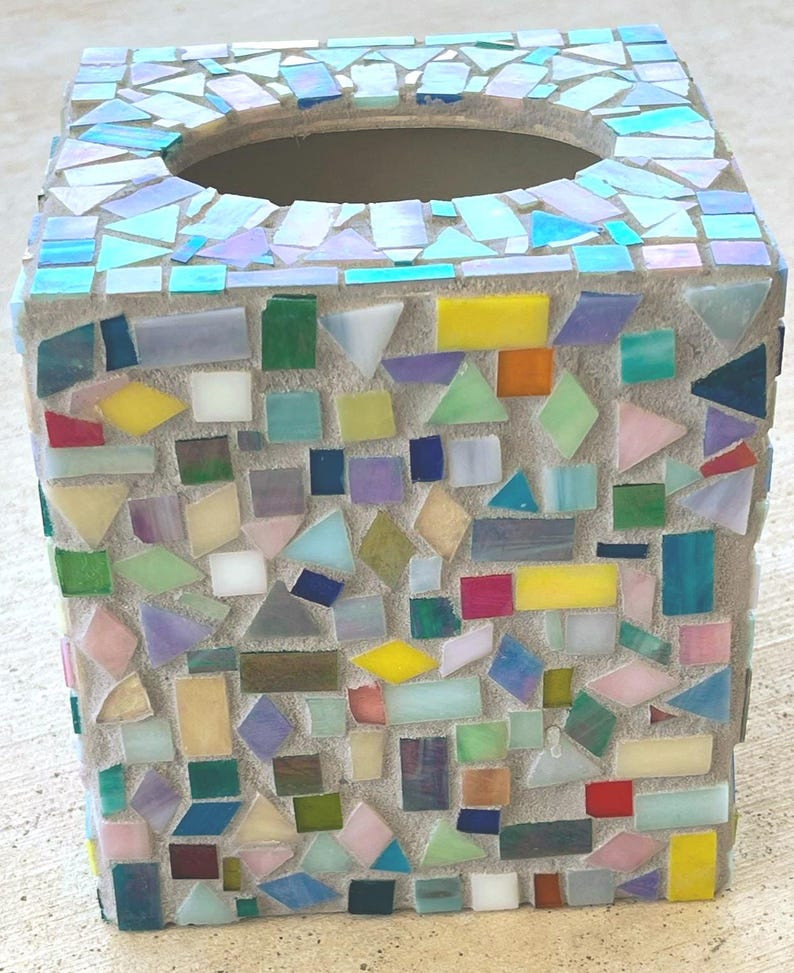 May include: A square tissue box cover decorated with a mosaic of multicolored glass tiles in shades of blue, yellow, green, pink, and orange. The tiles are set into a light gray grout, creating a textured surface with a circular opening on top.