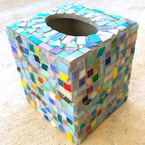 May include: A square tissue box cover featuring a mosaic design made of small, multi-colored glass tiles in shades of blue, green, yellow, red, and iridescent white, all set into a light gray grout base with a circular opening on the top surface.