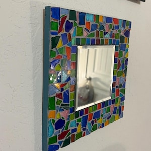 May include: A square wall mirror featuring a frame decorated with a mosaic of multi-colored glass shards in shades of blue, green, red, orange, and pink, set in gray grout. The mirror has a beveled edge and a solid blue painted side border.