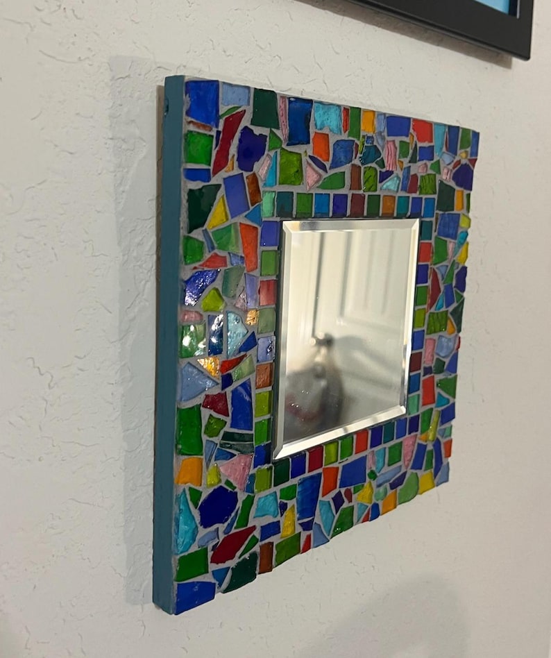 May include: A square wall mirror featuring a frame decorated with a mosaic of multi-colored glass shards in shades of blue, green, red, orange, and pink, set in gray grout. The mirror has a beveled edge and a solid blue painted side border.