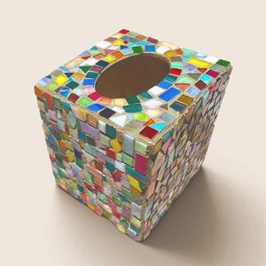 May include: A square tissue box cover featuring a multi-colored mosaic design made of small, irregular glass and ceramic tiles in shades of red, blue, green, yellow, and white, with a central oval opening on the top surface for dispensing tissues.