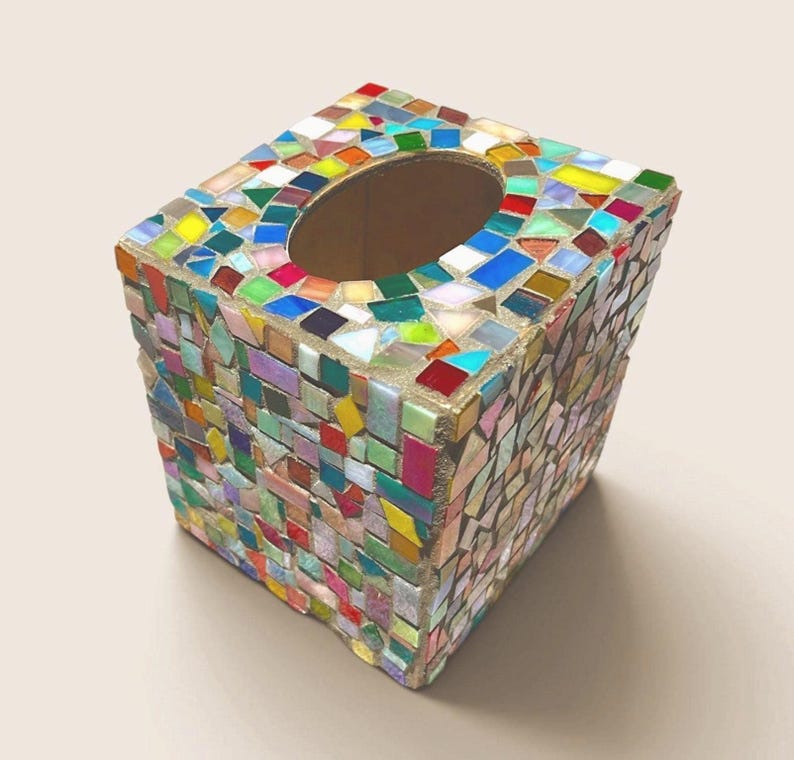 May include: A square tissue box cover featuring a multi-colored mosaic design made of small, irregular glass and ceramic tiles in shades of red, blue, green, yellow, and white, with a central oval opening on the top surface for dispensing tissues.